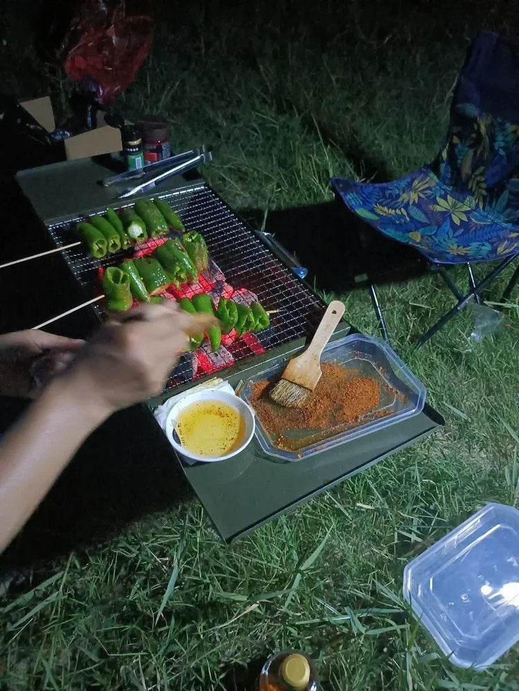 Outdoor barbeque grilling green peppers on skewers over charcoal, with spices and oil on the side.