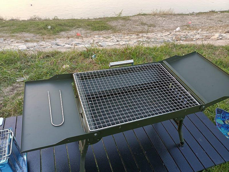 Outdoor portable metal grill with folding side shelves and mesh cooking surface by a riverside.