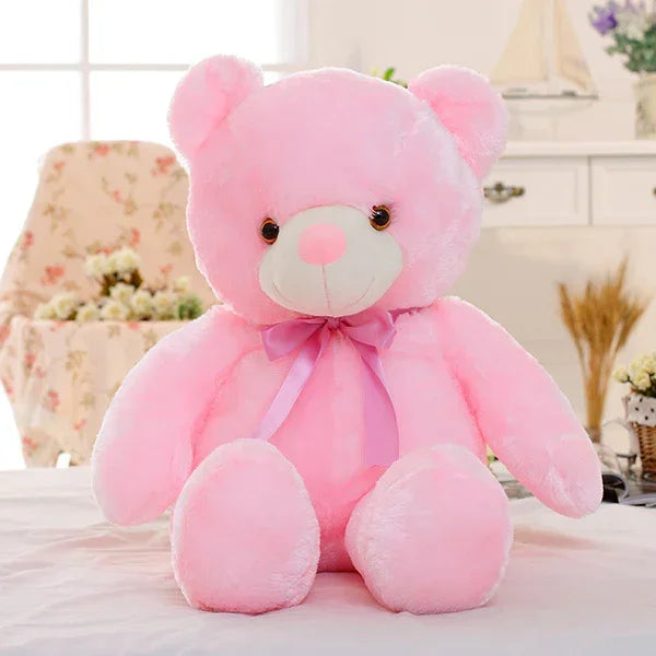 Large plush pink teddy bear with a bow, soft fur, sitting on a bed in a cozy room
