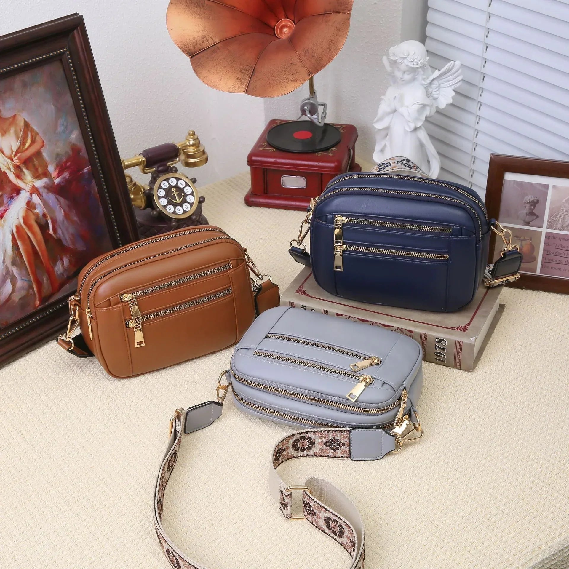 Three small crossbody bags in brown, navy blue, and light gray vegan leather, with gold zippers and adjustable straps, displayed on a neutral surface.