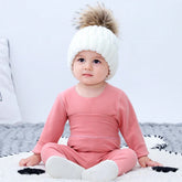 Baby in soft pink long sleeve romper and white knit beanie with faux fur pompom, sitting indoors