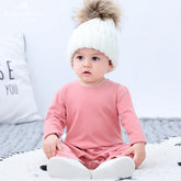 Baby wearing a soft pink long sleeve romper and white knit beanie with fur pompom, sitting indoors