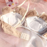 Beige and white padded bra with faux fur trim, adjustable straps, and hook closure, laid on pink tulle fabric.
