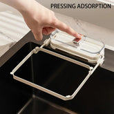 Hand pressing clear plastic sink-mounted trash bag holder with rectangular frame, modern kitchen accessory.
