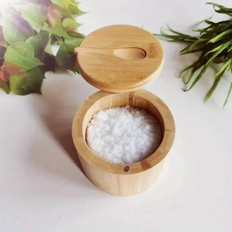 Round bamboo salt cellar with white coarse salt on white surface, green leaves nearby