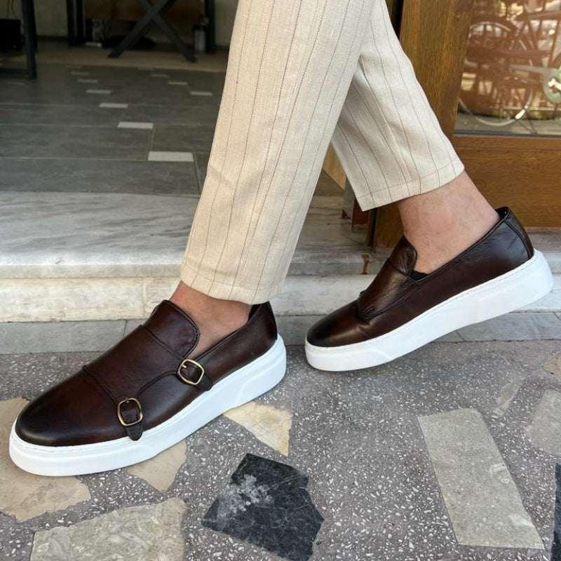 Men’s brown leather slip-on sneakers with buckles and thick white soles, paired with beige striped pants.