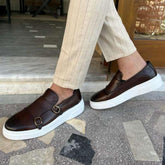 Men’s brown leather slip-on sneakers with buckles and thick white soles, paired with beige striped pants.
