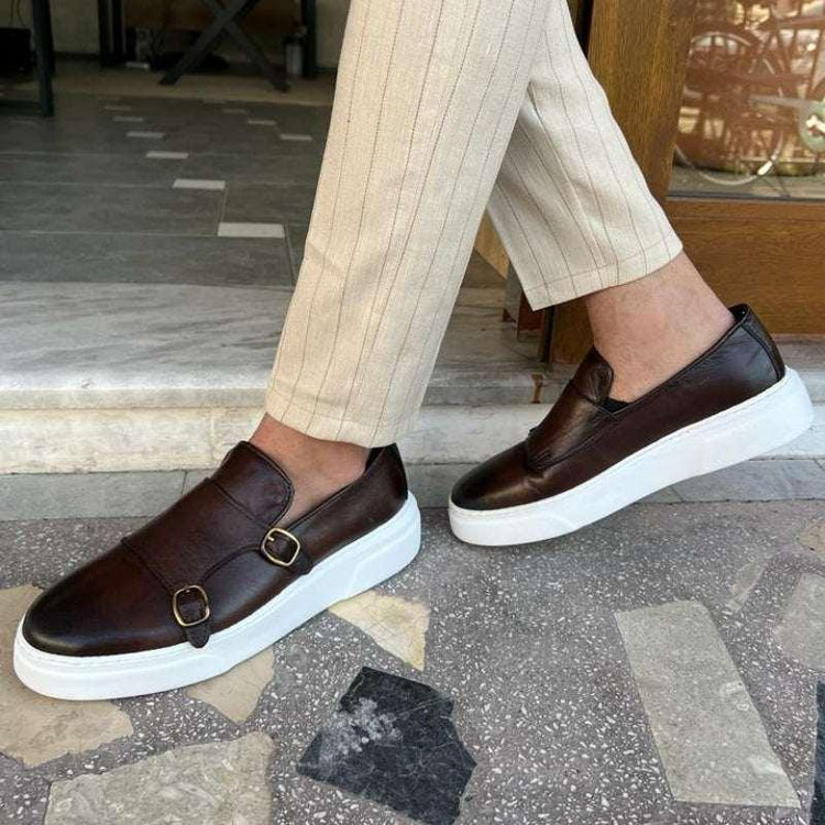Men’s brown leather slip-on sneakers with buckles and thick white soles, paired with beige striped pants.