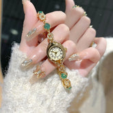 Gold bracelet watch with green and crystal accents, paired with glittery acrylic nails, fuzzy cream sweater
