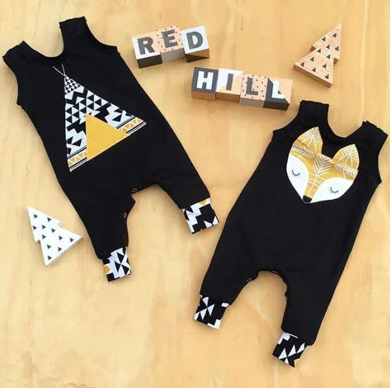 Two black cotton baby rompers with geometric patterns, one featuring a fox face and the other a yellow teepee, both with patterned cuffs, laid flat on a wooden surface.