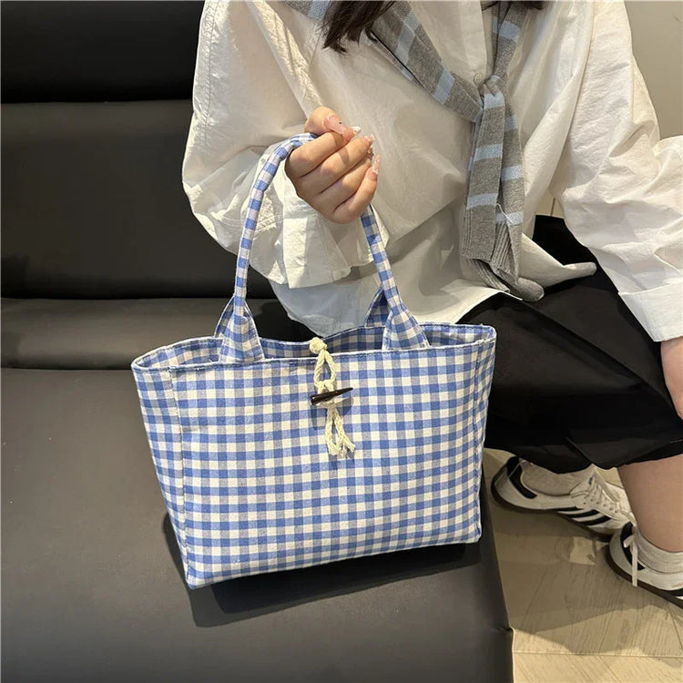 Blue and white checkered tote bag with rope toggle, held by person in oversized white shirt and black shorts.