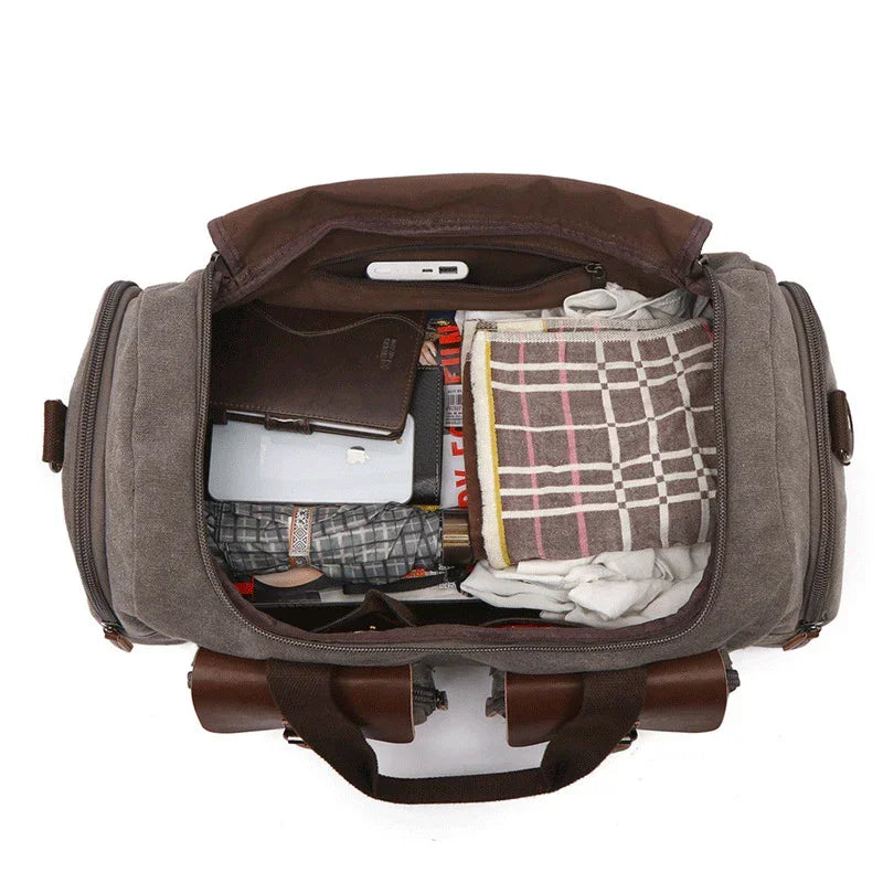 Open canvas duffel bag with brown leather accents, packed with folded plaid clothing and accessories.