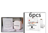 Compact white portable thermal printer with charging cable and six rolls of sticker printer paper.