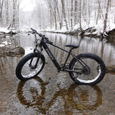 Black fat tire mountain bike with snow-covered tires parked in a snowy forest creek