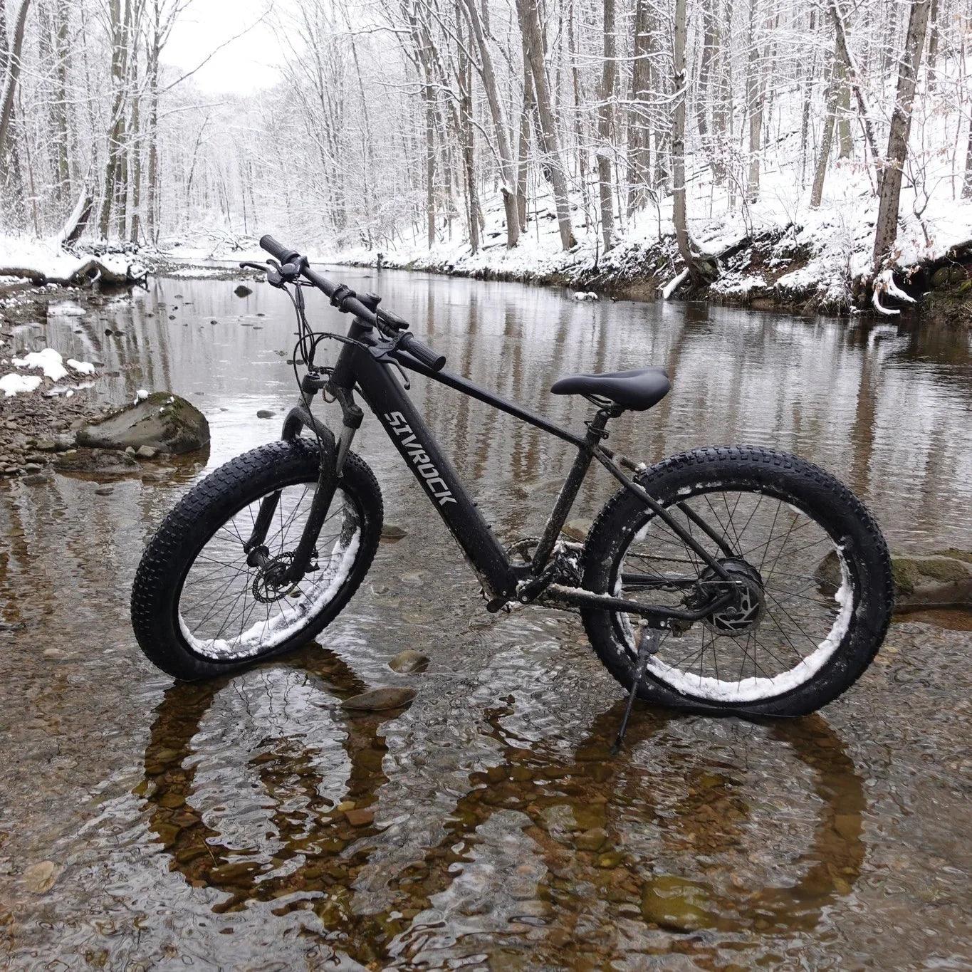 Black fat tire mountain bike with snow-covered tires parked in a snowy forest creek