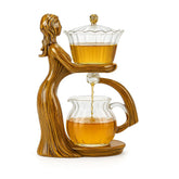 Artistic glass tea set with amber liquid, featuring a woman-shaped wooden stand and elegant curved design.