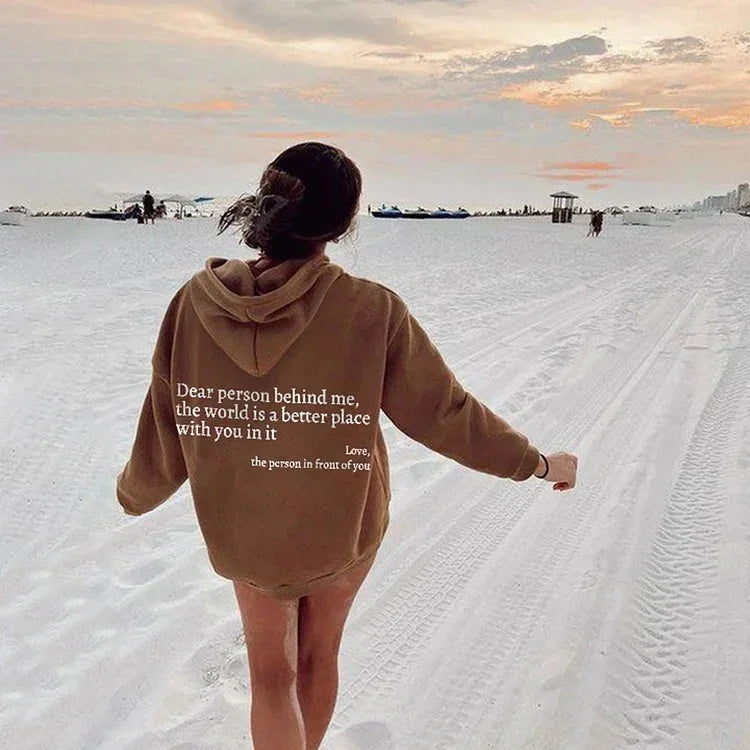 Woman in oversized brown hoodie with white quote, relaxed fit streetwear, walking on sandy beach at sunset.