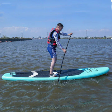 Man paddleboarding wearing blue patterned athletic shirt, blue shorts, and red life vest