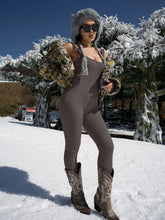 Woman in taupe ribbed fitted jumpsuit, floral puffer jacket, faux fur hat, and cowboy boots standing on snow.