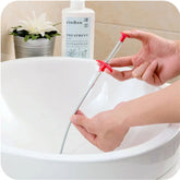 Hands using a flexible metal drain cleaning tool with red handle in a white sink.