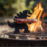 Metal dinosaur-shaped grill tool holding sausage on barbecue with open flame
