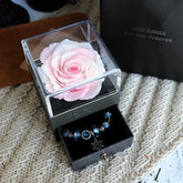 Pink preserved rose in a clear display box with a drawer holding a blue beaded bracelet.