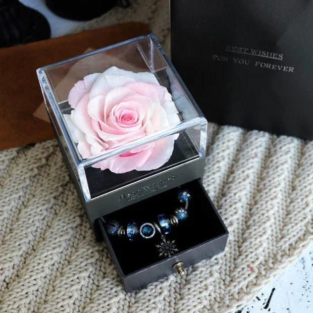 Pink preserved rose in a clear display box with a drawer holding a blue beaded bracelet.