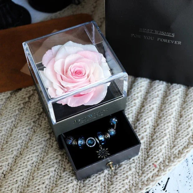 Pink preserved rose in a clear display box with a drawer holding a blue beaded bracelet.
