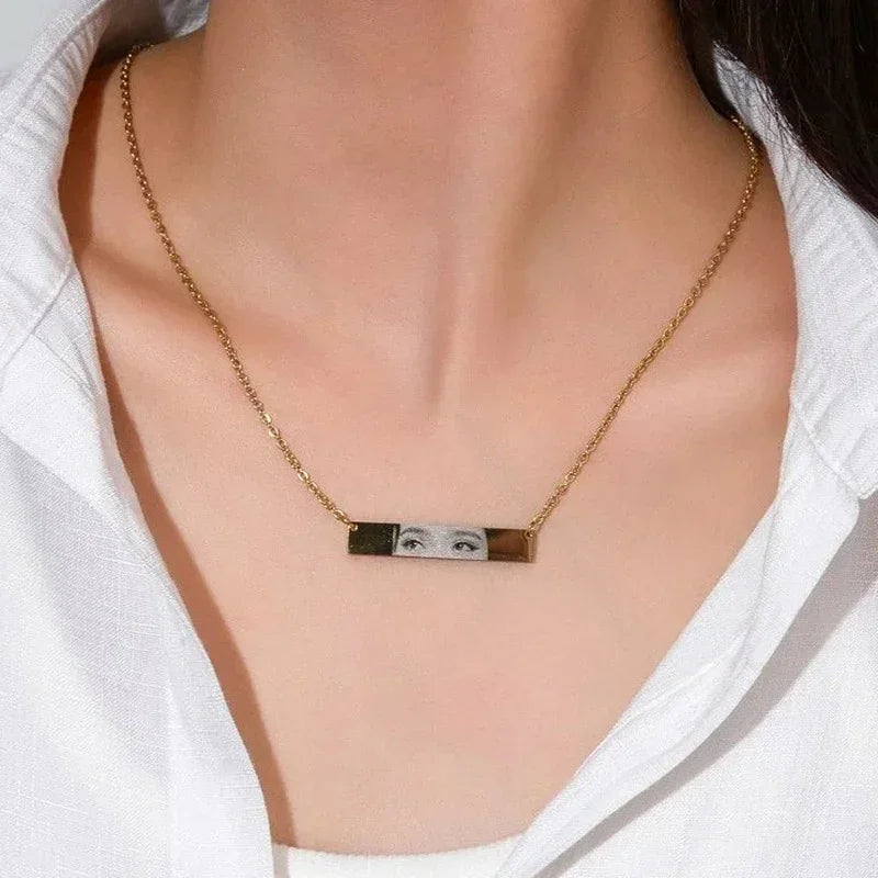 Gold bar necklace with engraved eye graphic, worn over a white collared shirt
