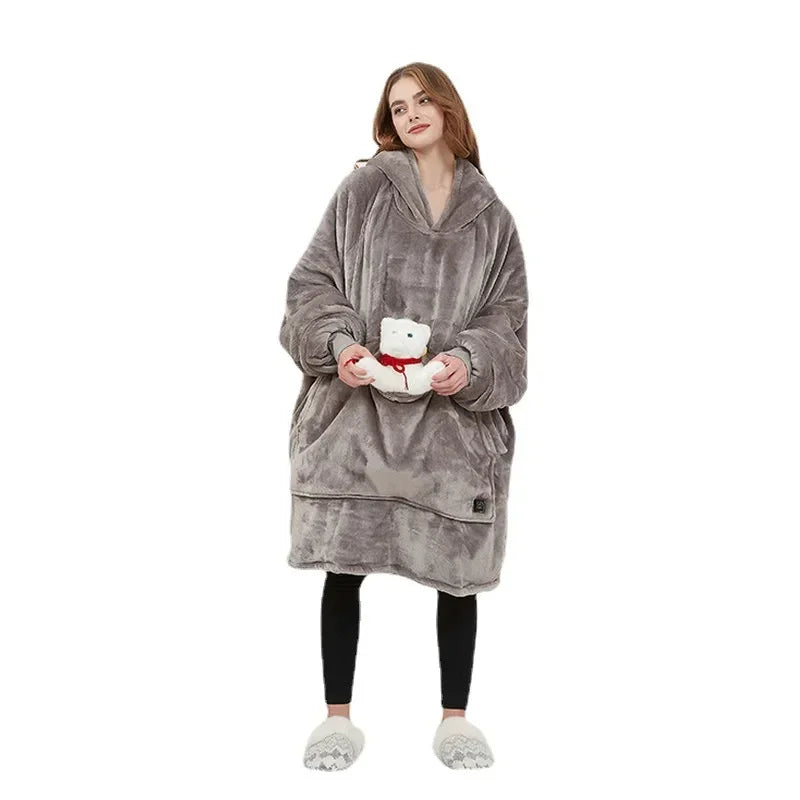 Woman wearing oversized grey fleece hoodie blanket, black leggings, and slippers, holding plush toy