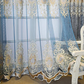 Sheer blue curtain with gold embroidered floral patterns and scalloped edges, draped in front of a window.