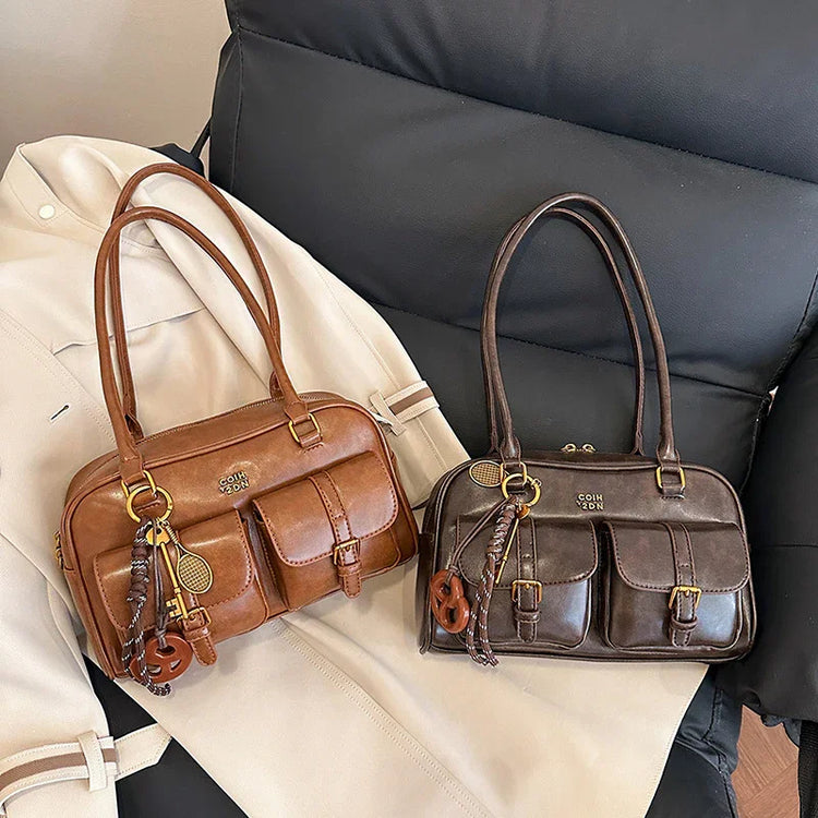 Two leather shoulder bags, one brown and one dark brown, with front pockets and gold hardware, displayed on a pale coat and black chair.