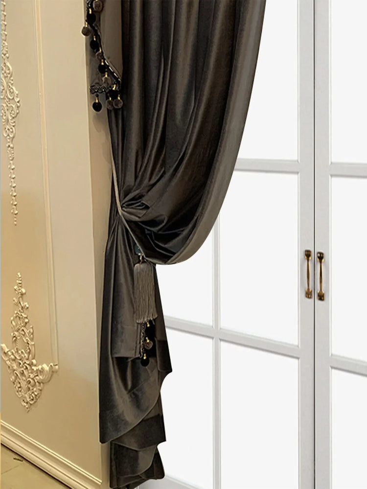 Dark gray velvet curtain with tassel and pom-pom trim, draped by a window