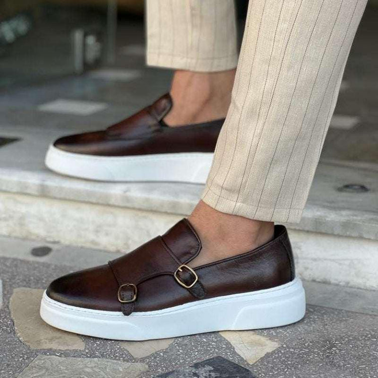 Brown leather slip-on sneakers with white soles and gold buckles, paired with beige pinstripe pants