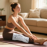 Woman in taupe sports bra and leggings, wearing a white compression knee sleeve, stretching on a yoga mat in a modern living room.