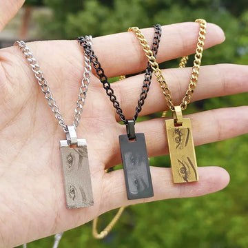 Three streetwear necklaces with rectangular pendants featuring engraved eye designs in silver, black, and gold, displayed on a hand outdoors.
