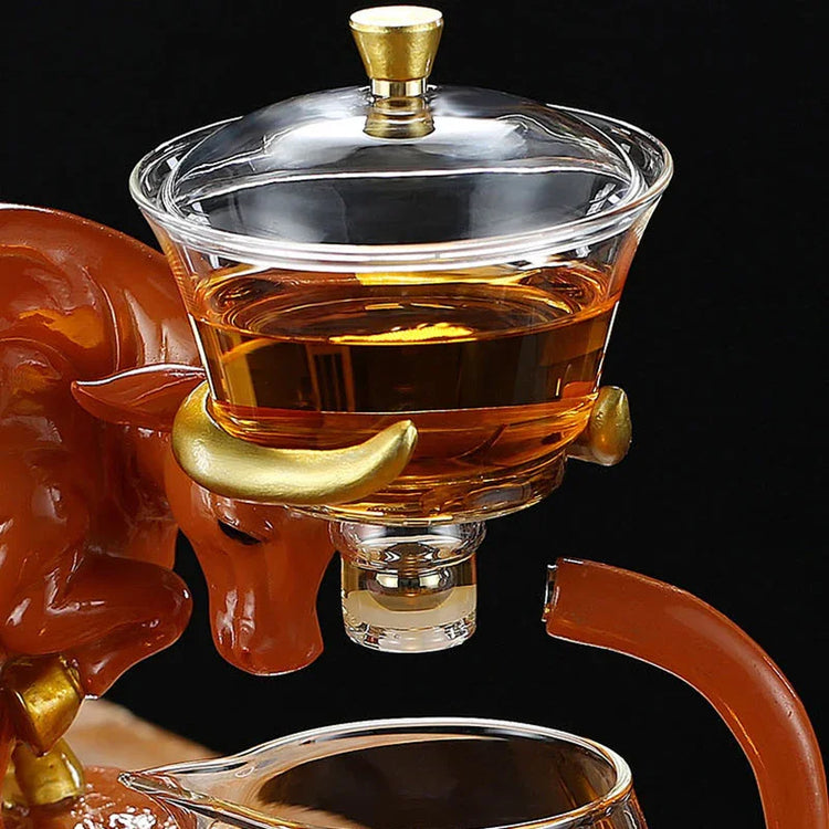 Clear glass teapot with gold accents shaped like a bull, containing amber tea, on black background.