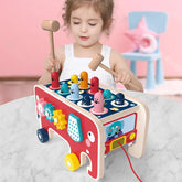 Toddler in a white sleeveless dress playing with a colorful wooden whack-a-mole toy at a table.