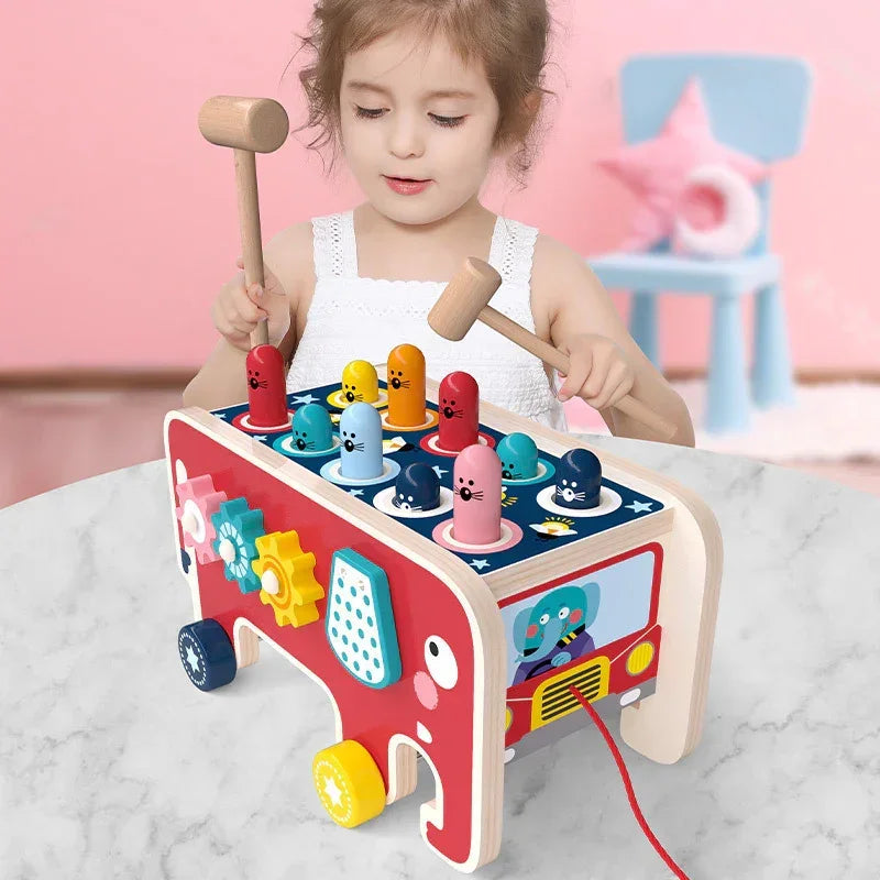 Toddler in a white sleeveless dress playing with a colorful wooden whack-a-mole toy at a table.