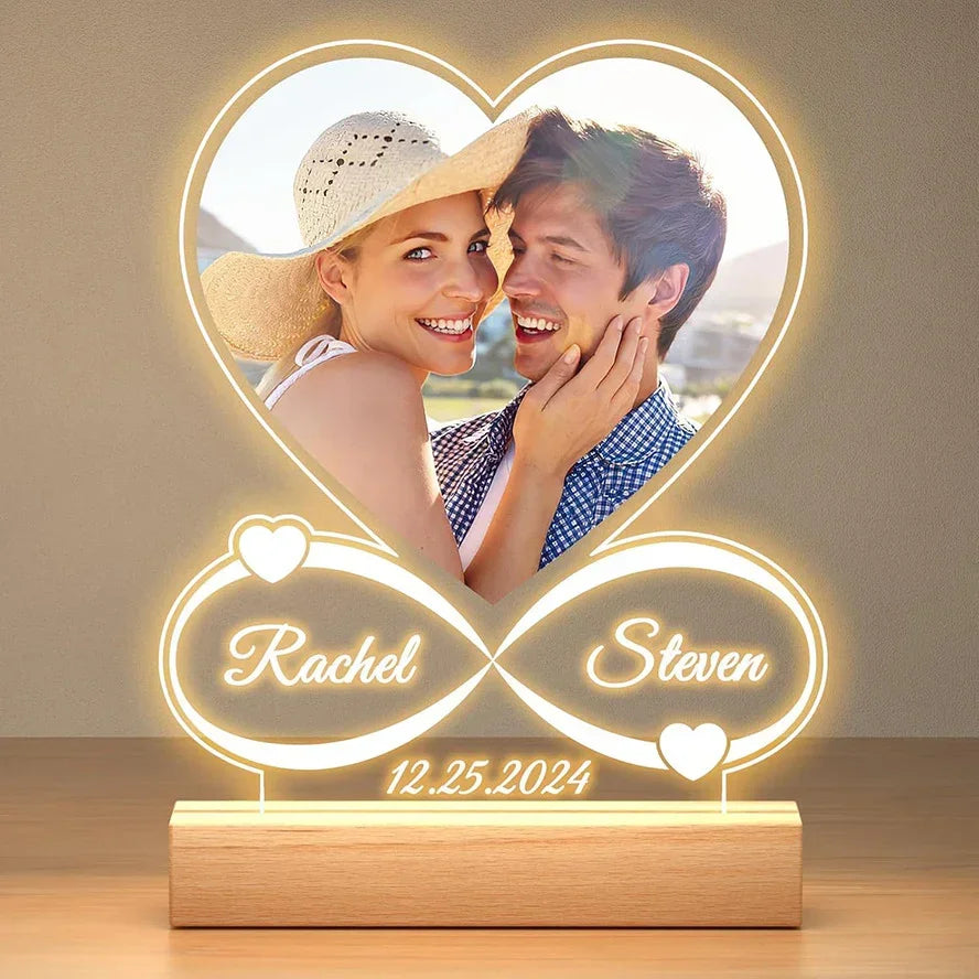 Personalized LED acrylic heart-shaped night light with engraved names, infinity design, and date, displayed on a wooden base.