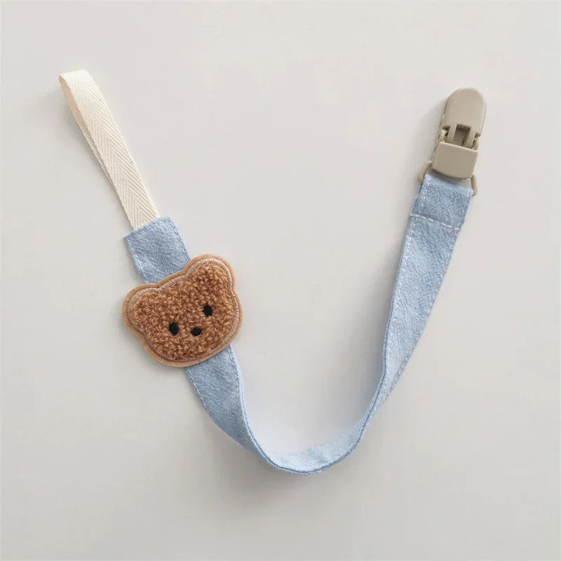 Blue fabric pacifier clip with beige teddy bear patch and cream loop, soft and cute design