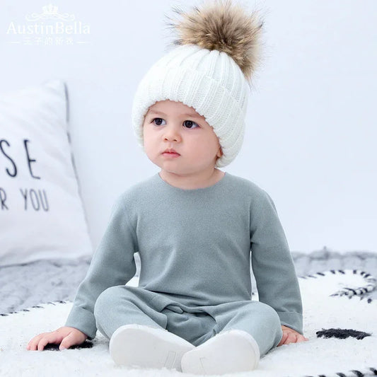 Baby in light grey long-sleeve outfit and white knit beanie with faux fur pom, relaxed fit.
