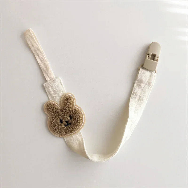 Cream cotton pacifier clip with beige bear patch and plastic clasp, minimalist style.