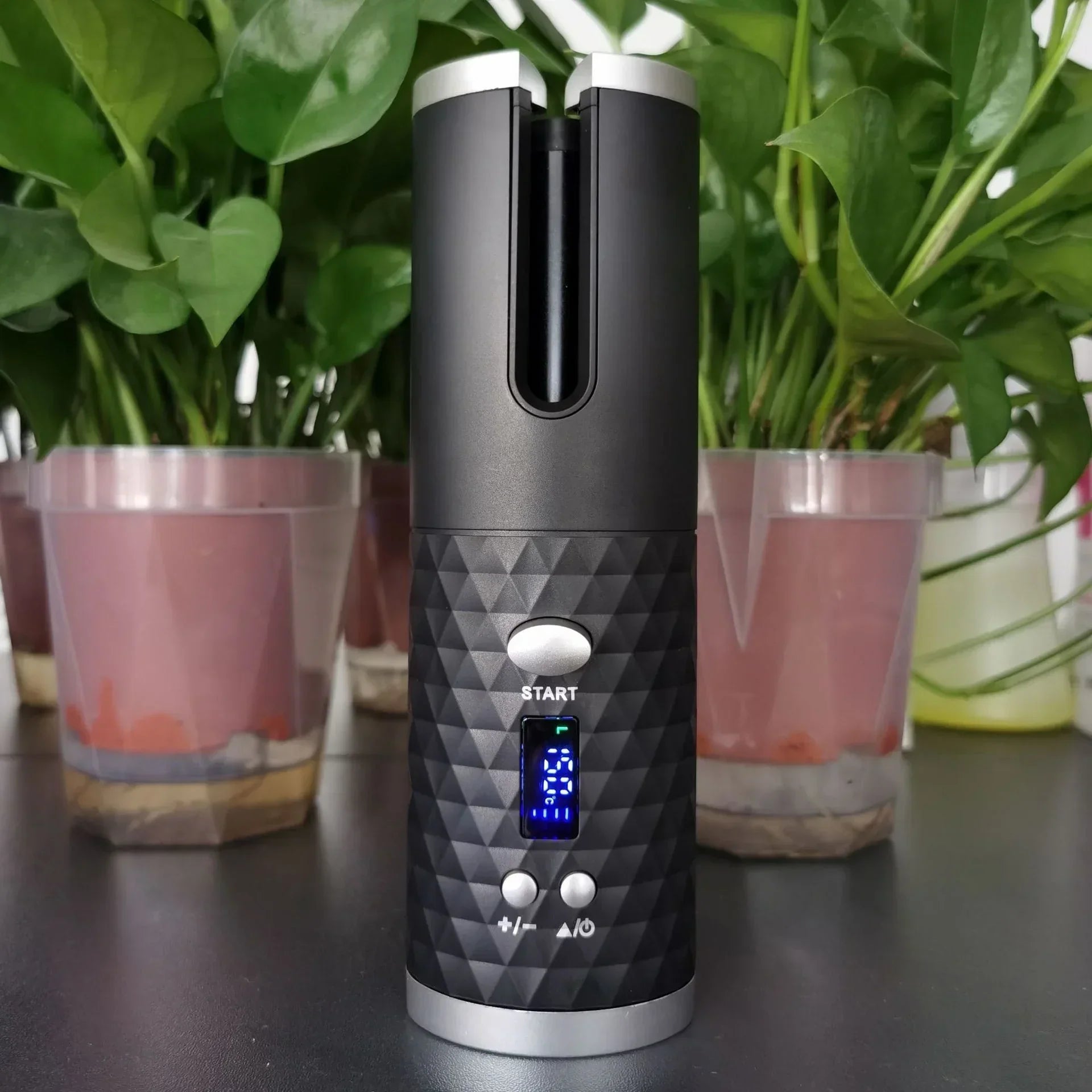 Black portable automatic hair curler with diamond-textured grip and digital display, on a table with green plants in the background.