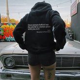 Woman in a loose black hoodie with positive quote, paired with black denim shorts, standing by a vintage car, urban street style.