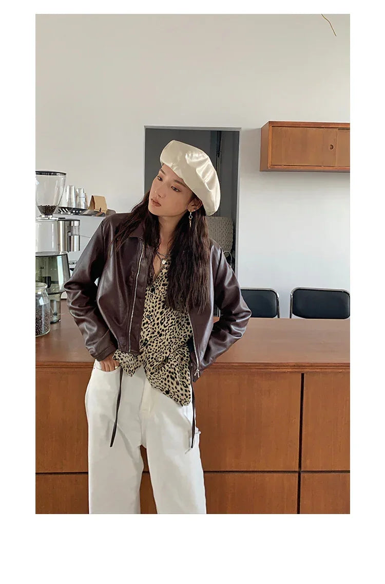 Woman in a brown faux leather jacket, leopard print blouse, white wide-leg pants, and cream beret