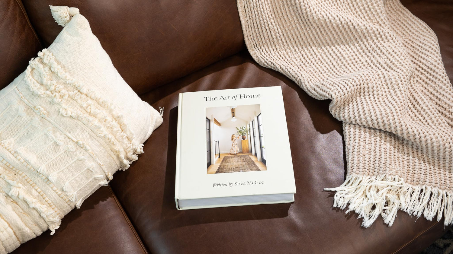 The Art of Home hardcover book by Shea McGee on brown leather sofa with cream textured pillow and blanket