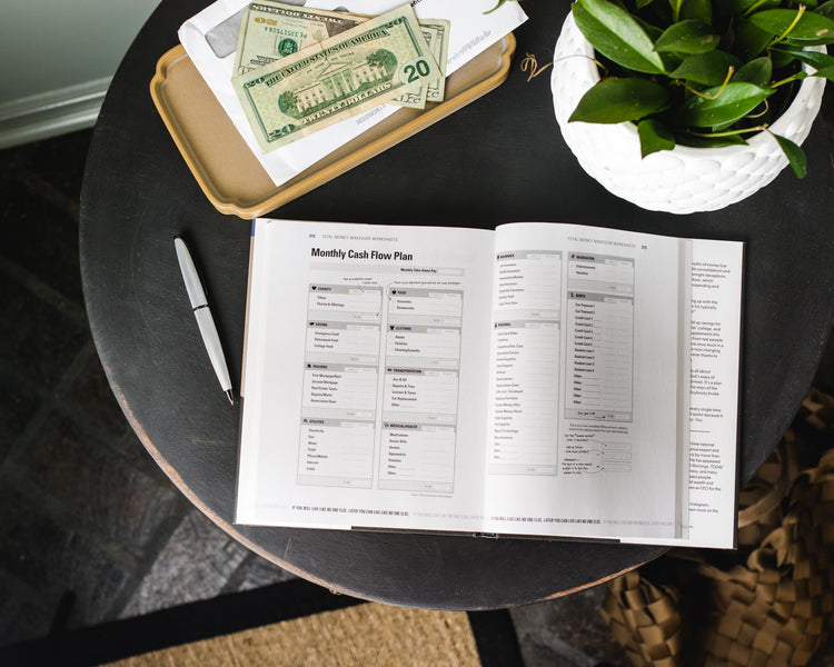 Open Total Money Makeover book showing Monthly Cash Flow Plan on table with US dollar bills and plant