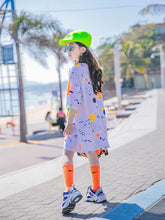 Girl wearing oversized pastel streetwear T-shirt dress with abstract print, bright neon cap, orange socks, and chunky sneakers outdoors.