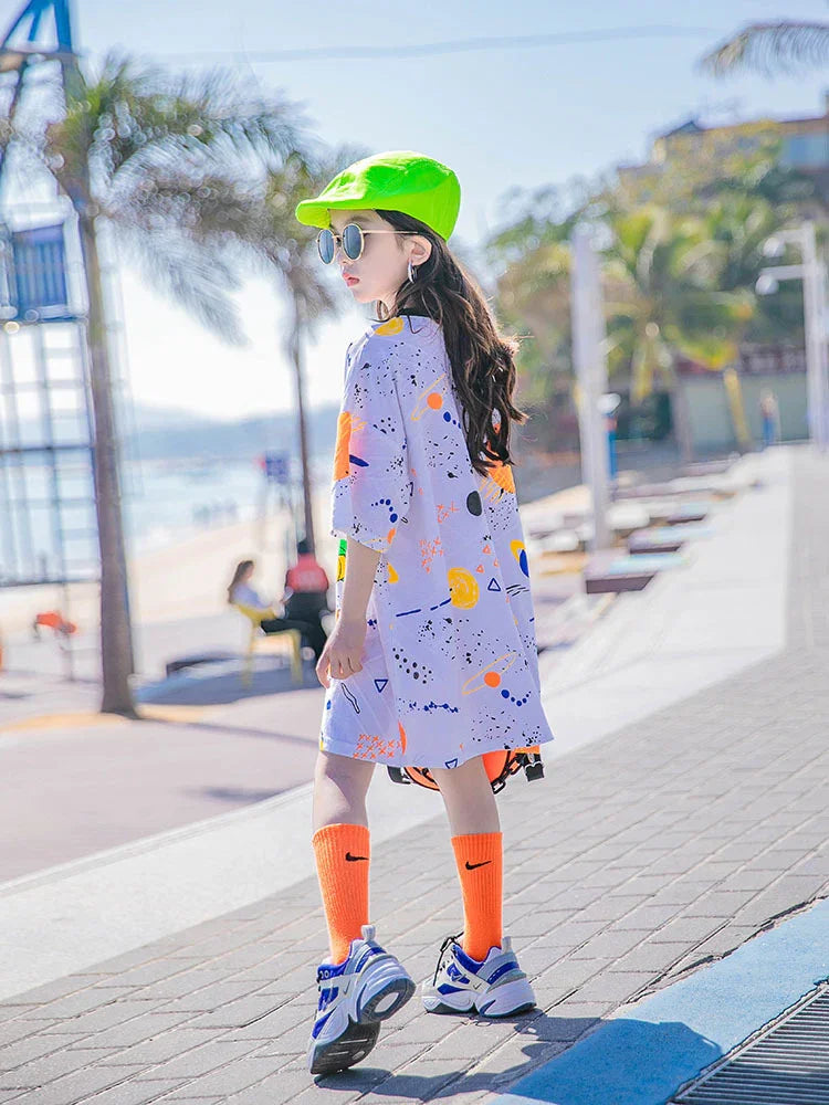 Girl wearing oversized pastel streetwear T-shirt dress with abstract print, bright neon cap, orange socks, and chunky sneakers outdoors.